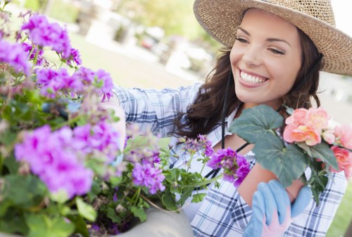 A close-up view of a woman gardening outdoors in a lush, well-maintained garden. She is smiling and wearing a wide-brimmed straw hat, blue gardening gloves, and a light blue checkered shirt. In front of her are vibrant pink, purple, and peach-colored flowering plants, with a background of green foliage and trees indicating a sunny, pleasant day. The scene reflects a landscaped outdoor space with neatly arranged flower beds, grass, and garden elements suggestive of professional gardening services in a residential garden near Mayfair, London, with natural tones of greenery, floral colours, and well-kept foliage. The woman appears to be tending to or planting the flowers, contributing to the garden's cultivated appearance.
